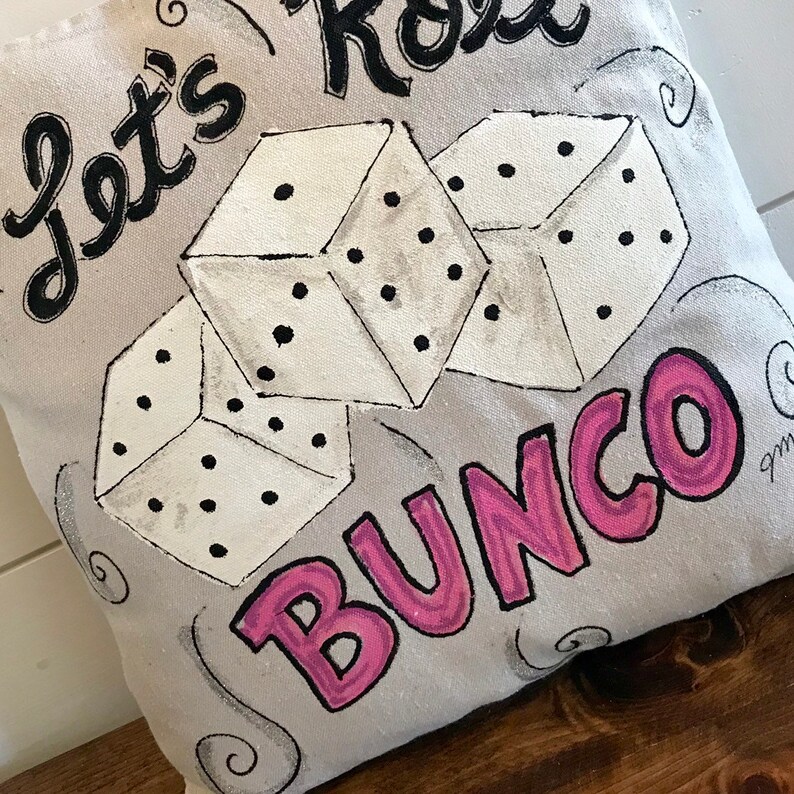Peut inclure: Un coussin en toile grise avec les mots "Let's Roll Bunco" en lettres noires et roses. Trois d&eacute;s blancs avec des points noirs sont peints sur le coussin. Le coussin est pos&eacute; sur une surface en bois.
