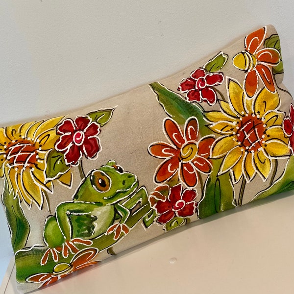 May include: A decorative pillow with a green frog sitting on a branch with yellow, red, and orange flowers. The pillow is made of a light brown fabric.
