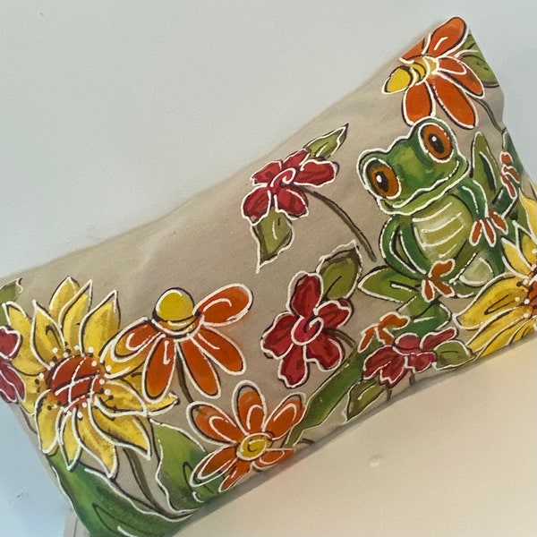 May include: A beige linen pillow with a hand-painted design featuring a green frog, sunflowers, and red, orange, and yellow flowers.