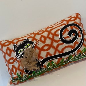 May include: A rectangular throw pillow featuring an orange and white geometric pattern. A black cat illustration with green eyes and a long, curled tail is painted on the front, accented by a textured brown burlap bow at the cat's neck.