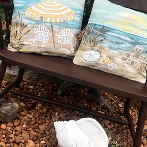 May include: Two decorative throw pillows with a beach theme. One pillow features a yellow and white striped umbrella with two beach chairs on a sandy beach. The other pillow features a seashell, a sand dollar, and a seascape with a sandy beach and a blue ocean.