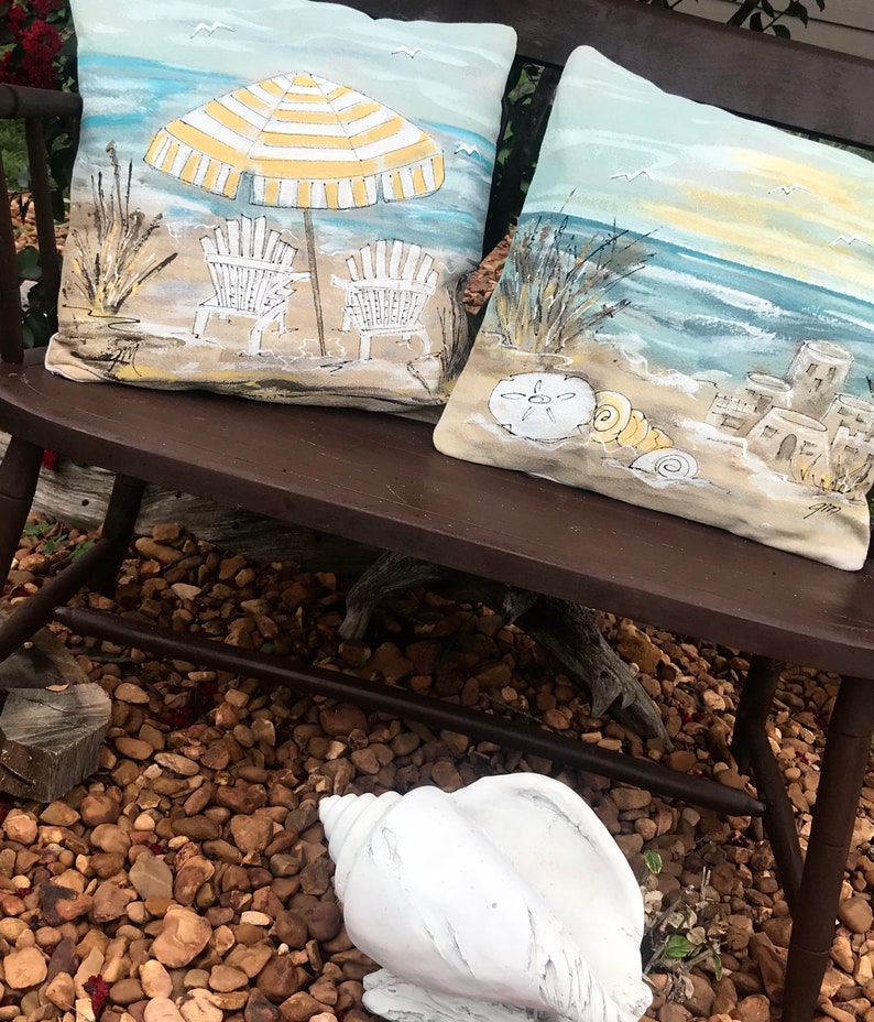 May include: Two decorative throw pillows with a beach theme. One pillow features a yellow and white striped umbrella with two beach chairs on a sandy beach. The other pillow features a seashell, a sand dollar, and a seascape with a sandy beach and a blue ocean.