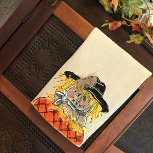 May include: A hand-painted kitchen towel featuring a whimsical scarecrow with a black hat, orange and white plaid shirt, and a blue bow tie. The scarecrow has a friendly face with yellow hair and a stitched smile.