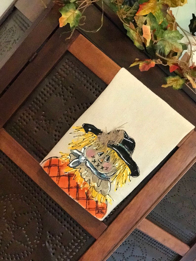 May include: A hand-painted kitchen towel featuring a whimsical scarecrow with a black hat, orange and white plaid shirt, and a blue bow tie. The scarecrow has a friendly face with yellow hair and a stitched smile.