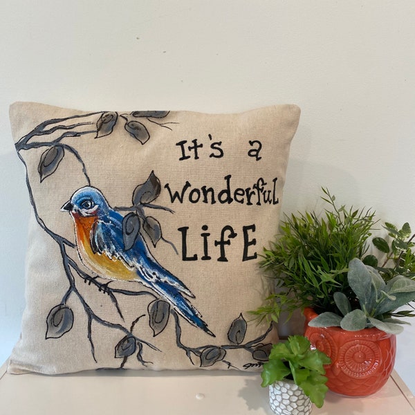 May include: A beige throw pillow with a bluebird perched on a branch with gray leaves. The pillow has the text "It's a wonderful life" printed on it.
