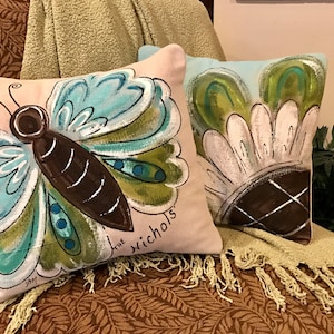 May include: Two decorative throw pillows with hand-painted designs. One pillow features a blue and green butterfly with brown accents and the text "the nichols". The other pillow features a white flower with green leaves and a brown center.