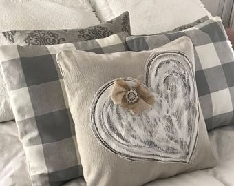 May include: A white and grey checked pillow with a beige pillow featuring a white painted heart with a burlap bow on top. The pillows are on a white bed with a white headboard.