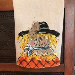 May include: A hand-painted kitchen towel featuring a whimsical scarecrow with a black hat, yellow straw hair, and a blue scarf. The scarecrow is wearing a brown shirt and has a pumpkin-like face with a stitched smile. The background is a checkered orange and brown pattern.
