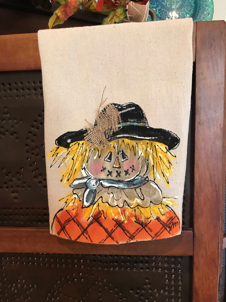 May include: A hand-painted kitchen towel featuring a whimsical scarecrow with a black hat, yellow straw hair, and a blue scarf. The scarecrow is wearing a brown shirt and has a pumpkin-like face with a stitched smile. The background is a checkered orange and brown pattern.