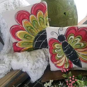 K&ouml;nnte beinhalten: Zwei dekorative Kissen mit handgemalten Designs. Ein Kissen zeigt ein rotes, gelbes und gr&uuml;nes Blumendesign. Das andere Kissen zeigt einen schwarzen Schmetterling mit roten, gelben und gr&uuml;nen Fl&uuml;geln.