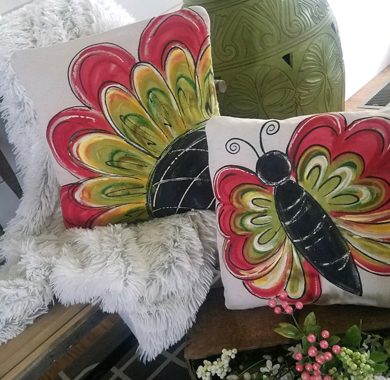 K&ouml;nnte beinhalten: Zwei dekorative Kissen mit handgemalten Designs. Ein Kissen zeigt ein rotes, gelbes und gr&uuml;nes Blumendesign. Das andere Kissen zeigt einen schwarzen Schmetterling mit roten, gelben und gr&uuml;nen Fl&uuml;geln.