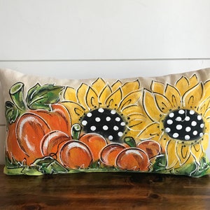 May include: A decorative pillow with a hand-painted design featuring sunflowers and pumpkins. The sunflowers have black and white polka dot centers and the pumpkins are orange with green leaves.