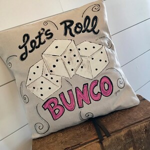 Peut inclure: Un coussin carr&eacute; beige avec les mots "Let's Roll Bunco" en noir et rose. Trois d&eacute;s blancs avec des points noirs sont repr&eacute;sent&eacute;s au centre. Le coussin repose sur une surface en bois.