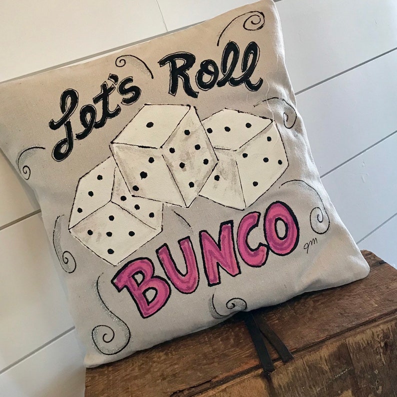 Peut inclure: Un coussin carr&eacute; beige avec les mots "Let's Roll Bunco" en noir et rose. Trois d&eacute;s blancs avec des points noirs sont repr&eacute;sent&eacute;s au centre. Le coussin repose sur une surface en bois.