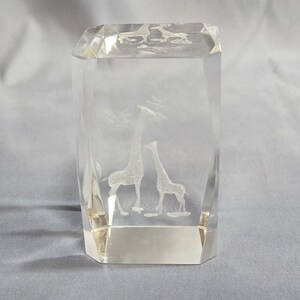 May include: A clear, rectangular crystal block featuring an etched design of two giraffes. The giraffes are side-by-side, with one drinking water. The block has a faceted top and base, and is approximately 7 cm tall.