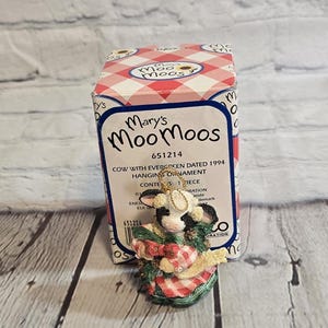 May include: A Mary's MooMoos cow figurine with a red and white checkered dress and a green base. The figurine is a hanging ornament and is packaged in a box with the text "Mary's MooMoos".