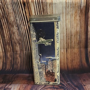 May include: Rectangular metal tin with a hinged lid. The tin features a nighttime cityscape with a biplane flying over skyscrapers. Text on the side reads "Authentic...Quality...letter". The tin is silver and the background is a dark blue.