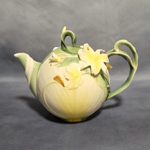 May include: A ceramic teapot featuring a rounded body with a pale yellow and green gradient finish. The teapot is adorned with three-dimensional sculpted white and yellow lily flowers and green leaves. It includes a curved handle and a lid.