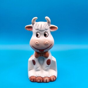 May include: A white ceramic cow figurine with brown spots and a red bow tie. The cow is sitting with its head tilted to the side and has a playful expression.
