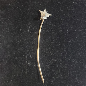 May include: A silver-toned metal pin with a star-shaped design. The pin is long and slender, with a curved stem.