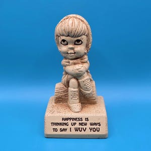 May include: A beige figurine of a seated child with a scarf, holding their arms around themselves. The base reads "Happiness is thinking up new ways to say I wuv you."