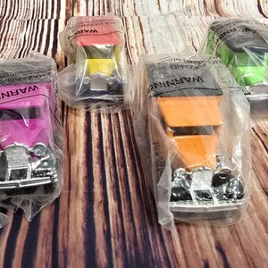 May include: Four vintage toy cars in different colors, including pink, orange, green, and yellow, are displayed in plastic packaging. Each car has a black grill and headlights. The packaging includes the word "WARNING" printed on it.