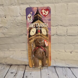 May include: A brown Britannia the Bear Beanie Baby toy in its original packaging. The packaging features an image of Big Ben and the text "Britannia the Bear". The bear has a red ribbon and a Union Jack on its chest.
