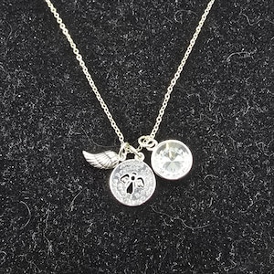 May include: Silver necklace featuring a wing charm, a circular pendant with an angel motif, and a clear crystal pendant. The necklace is displayed against a black backdrop.