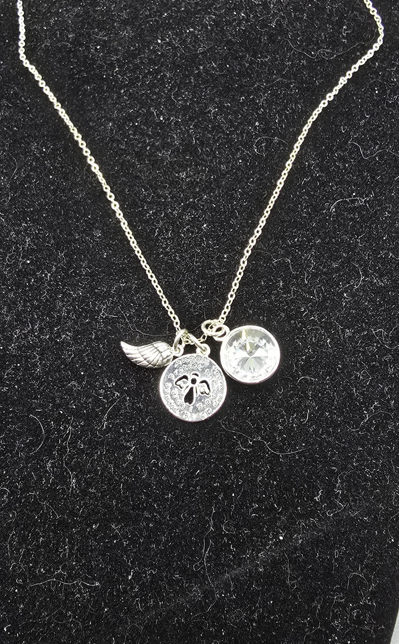 May include: Silver necklace featuring a wing charm, a circular pendant with an angel motif, and a clear crystal pendant. The necklace is displayed against a black backdrop.