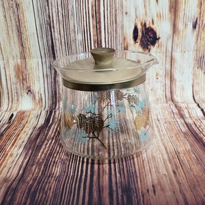 May include: A clear glass pitcher with a gold lid and a pine cone design. The pitcher has a wide base and a narrow spout. The pine cones are brown and gold, and the pine needles are green.