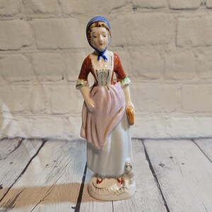 May include: A porcelain figurine of a woman wearing a red and white dress with a pink apron and a blue bonnet. She is holding a book and has a small white dog at her feet.