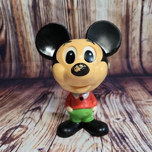 May include: A vintage Mickey Mouse figurine. The figure has a tan face, black ears, and a red shirt with green shorts. The figurine is standing on a wooden surface.