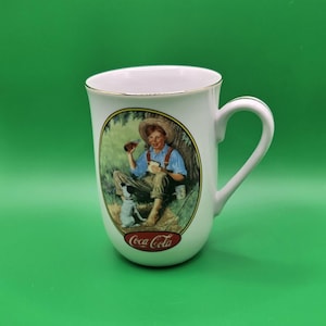 May include: White ceramic Coca-Cola mug with a gold rim and handle. The mug features a vintage illustration of a boy and dog enjoying a Coca-Cola. The Coca-Cola logo is at the bottom of the mug.