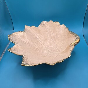 May include: A decorative leaf-shaped bowl with a cream-colored interior and gold-colored trim. The bowl has a textured surface and a detailed leaf vein pattern. The bowl is made of ceramic or a similar material.