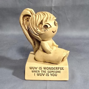 May include: A ceramic figurine of a girl with blonde hair in a ponytail, sitting with her legs crossed. The figurine is on a base with the text "WUV IS WONDERFUL WHEN THE SOMEONE I WUV IS YOU".
