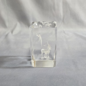 May include: A clear, rectangular crystal sculpture with an etched design of a deer, birds, and foliage. The artwork is inside the crystal block, creating a three-dimensional effect. The sculpture is displayed on a light grey surface.