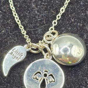 May include: Silver necklace featuring a chain and multiple charms. Charms include a round pendant with an angel design, a wing charm with the words "Little Angel", and a spherical charm. The necklace is displayed on a dark surface.