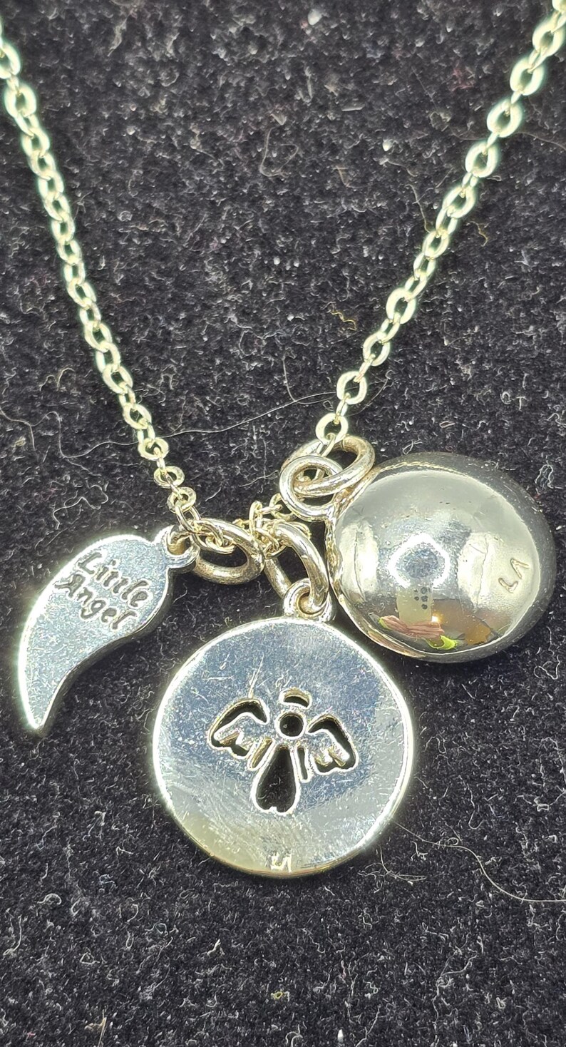May include: Silver necklace featuring a chain and multiple charms. Charms include a round pendant with an angel design, a wing charm with the words "Little Angel", and a spherical charm. The necklace is displayed on a dark surface.