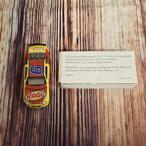 May include: A yellow, red, and blue toy race car with the number 6 on the side and the word "RAGU" printed on the front. The car is on a wooden surface. The box for the car is in the background and has the text "NASCAR® is a registered trademark of the National Association for Stock Car Auto Racing, Inc. Ragu®"