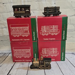 May include: A collection of miniature train cars and locomotives, including a passenger car labeled "Kemmerer WY. Express" and a timber car. The items are displayed on red boxes with train illustrations, suggesting a themed collectible.