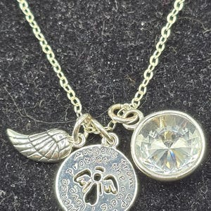 May include: Silver necklace featuring three charms. One is a wing, another a clear, round crystal, and the third a round pendant with the text "Angels protect the ones up above, please love us".