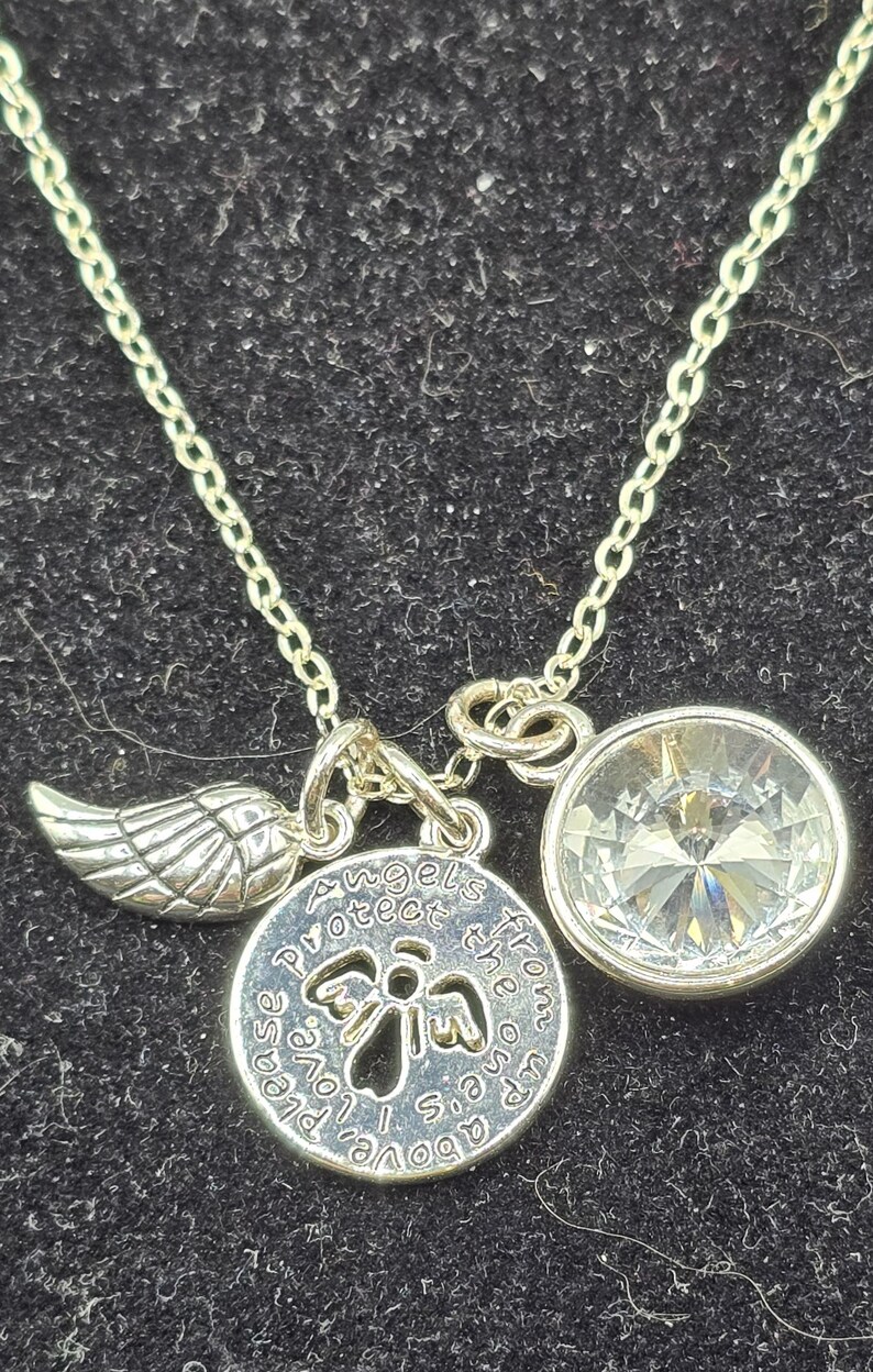 May include: Silver necklace featuring three charms. One is a wing, another a clear, round crystal, and the third a round pendant with the text "Angels protect the ones up above, please love us".