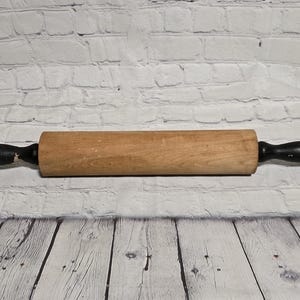May include: A wooden rolling pin featuring a natural light brown cylindrical barrel and two black painted handles on either end. The wood grain is visible across the center section, while the handles show signs of wear on the finish.