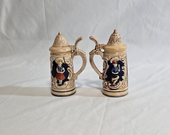 May include: Two beige ceramic salt and pepper shakers shaped like miniature beer steins. Each stein features a detailed relief design with a handle, lid, and a painted scene of a person in traditional clothing. The shakers are approximately 4 inches tall.