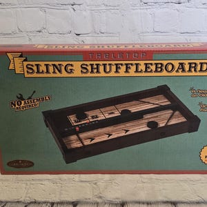 May include: A tabletop sling shuffleboard game in a red, green, and brown box. The box features the game's name and a picture of the game board. The box also states that no assembly is required and includes 3 pucks.