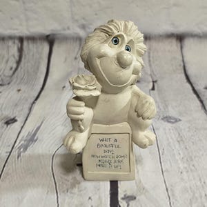 May include: A whimsical, off-white figurine of a smiling creature holding a rose. The figurine has blue eyes and a sign that reads "WHAT A BEAUTIFUL DAY! NOW WATCH SOME JERK MESS IT UP!"