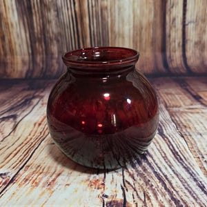 Peut inclure: Un vase rond en verre rouge foncé avec une large ouverture. Le vase a une finition lisse et brillante et repose sur une surface en bois. Le vase mesure environ 15 cm de diamètre et 13 cm de hauteur.
