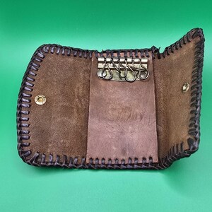 May include: Open brown leather key holder with a stitched border and metal key rings. The key holder is open, revealing the interior with key rings and a small leather flap. The background is a solid green.