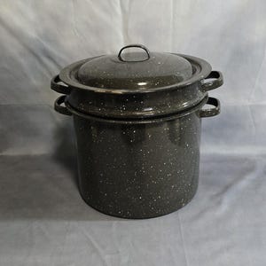 May include: A dark gray speckled enamelware pot with a lid. The pot has two side handles and a lid with a single handle. The pot is designed for cooking and has a classic, vintage aesthetic.