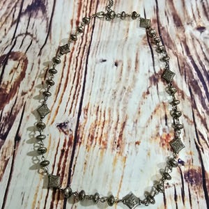 May include: Silver-toned necklace with a mix of round and square decorative elements. The necklace has a delicate chain and is displayed on a wooden surface. The square elements have a cross-like design.